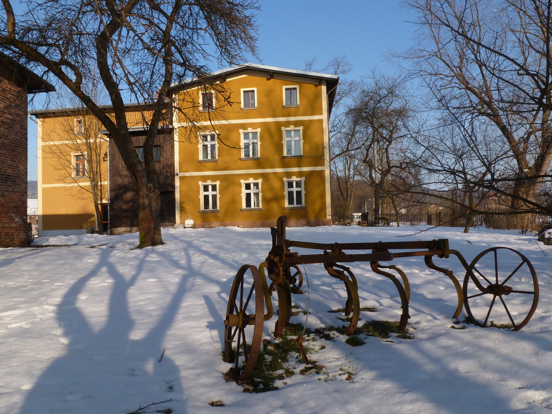 The yellow main house at Domstolat in the snow, with an old plough in the foreground